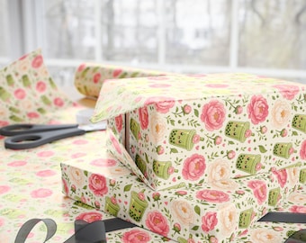 May include: Wrapping paper featuring a repeating pattern of pink and white peonies interspersed with green boba tea cups that have smiling faces. The paper is used to wrap rectangular boxes, with a pair of black scissors resting nearby.