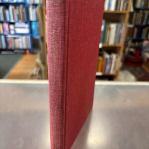 May include: A vintage, red hardcover book with gold lettering on the spine. The book's cover is a solid, deep red, and the edges show signs of wear. The background features bookshelves filled with other books, suggesting a library or bookstore setting.