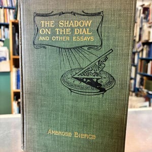 May include: A vintage book titled "The Shadow on the Dial and Other Essays" by Ambrose Bierce. The book has a textured green cover with gold lettering and a detailed illustration of a sundial. The book is standing upright.