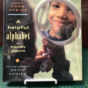 May include: A book cover with the title "A Helpful Alphabet of Friendly Objects" by John Updike. The cover features a photograph of a child looking through a circular mirror. The book is propped up on a white stand.