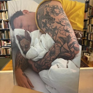 May include: A book cover showing a person and a small dog in white bedding. The person wears a yellow shirt and has arm tattoos. The dog, brown and white, is partially visible under the covers.