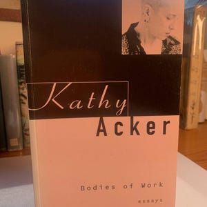Peut inclure: Une couverture de livre avec un fond rose et du texte noir. Le titre est "Kathy Acker" et le sous-titre est "Bodies of Work essays". Une photo en noir et blanc d'une femme aux cheveux courts se trouve dans le coin supérieur droit.