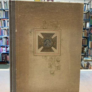 May include: A brown book with a gold embossed design on the front cover. The design features a cross with the words "Washington" and "1789" on it. The book is titled "The Washington Artillery, First Company".