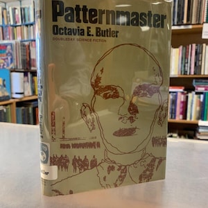 May include: A paperback book titled "Patternmaster" by Octavia E. Butler. The book cover features a brown and white illustration of a person's face with a crowd of people in the background. The book is a Doubleday Science Fiction publication.