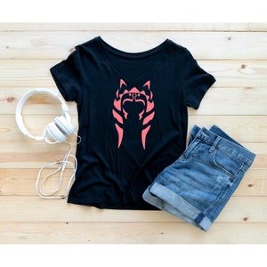 Op de afbeelding: Zwart T-shirt met een roze Ashoka Tano-symbool op de voorkant. Het shirt ligt plat op een houten oppervlak met een blauwe spijkerbroek en witte koptelefoon.