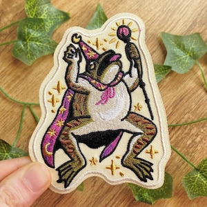 May include: Embroidered patch featuring a frog dressed as a wizard, wearing a purple and gold cape and hat. The frog is holding a staff with a glowing orb. The patch has a cream-colored background with gold star accents.