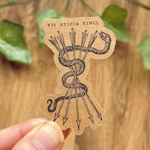 May include: A tan sticker featuring a black ink illustration of a snake coiled around a bundle of five arrows. The text VIS NESCIA VINCI is printed in black capital letters at the top. The sticker has a die-cut edge following the design shape.