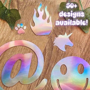 Könnte beinhalten: Eine Sammlung holografischer Aufkleber in verschiedenen Formen, darunter eine Flamme, ein Pfotenabdruck, ein Einhorn, ein @-Symbol und ein Smiley. Der Text "50+ designs available!" ist ebenfalls zu sehen. Die Aufkleber haben einen Regenbogen-Schimmer.