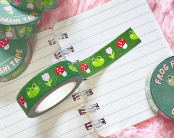 May include: Green washi tape rolls with a repeating pattern of smiling frogs, red mushrooms, and pink flowers. The tape is unrolled on a lined notebook. The words "Washi Tape" and "Frog Fun" are visible on the tape rolls.