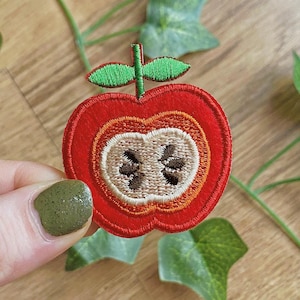 Puede incluir: Parche de manzana bordada con un diseño de sección transversal. El parche tiene un anillo exterior rojo, con detalles interiores naranjas y beige. El centro muestra las semillas en marrón. Las hojas y el tallo verdes completan el diseño.