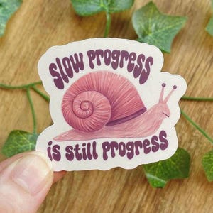 Può includere: Un adesivo raffigurante una lumaca rosa con un guscio a spirale. Il testo "slow progress is still progress" è scritto in viola con un carattere stilizzato. L'adesivo ha un bordo bianco ed è tenuto in mano.
