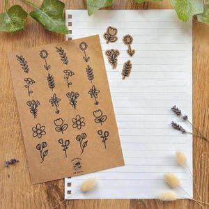 Könnte beinhalten: Ein rechteckiger Bogen braunes Kraftpapier mit einem Raster aus schwarzen floralen und botanischen Strichzeichnungen, einem kleinen Logo am unteren Rand mit der Aufschrift "LOKREAKISH CREATIONS" und mehreren einzelnen Blumenaufklebern in der Nähe.