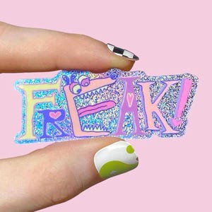 Peut inclure: Un autocollant scintillant avec le mot "FREAK!" dans une police de caractères de dessin animé. Les lettres sont de différentes couleurs, dont le rose, le violet et le jaune, avec un fond bleu pailleté. Le "E" est conçu pour ressembler à un monstre avec une longue langue.