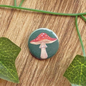 Könnte beinhalten: Ein runder Knopf mit einem roten Pilzdesign mit weißen Punkten auf dunklem grünen Hintergrund. Der Knopf befindet sich auf einer Holzoberfläche mit grünen Blättern und Ranken.