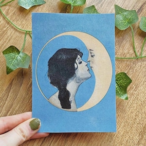 Op de afbeelding: Een blauwe kunstdruk met een vrouw en een halve maan in een cirkel. De vrouw heeft donker haar en kijkt naar de maan, die een gezicht heeft. De achtergrond is lichtblauw. De kunstdruk ligt op een houten oppervlak.