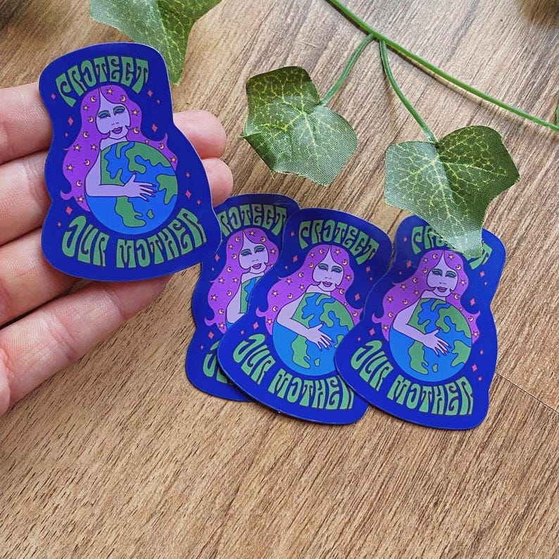 May include: A collection of blue stickers featuring a woman with purple hair embracing the Earth. The stickers have the text "PROTECT OUR MOTHER" in green, with a retro font. The stickers are on a wooden surface.