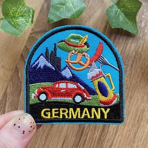 Könnte beinhalten: Gestickter Aufnäher mit blauem Hintergrund mit einem roten Auto, Bergen und einem Bierkrug. Der Aufnäher enthält eine Brezel, einen grünen Hut und das Wort "GERMANY" in Gelb.