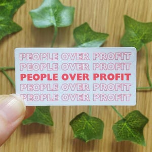 Könnte beinhalten: Ein weißer Aufkleber mit dem Text "PEOPLE OVER PROFIT" in einem sich wiederholenden Muster aus Rosa und Weiß. Der Aufkleber wird von einer Hand gehalten, mit grünen Blättern im Hintergrund.