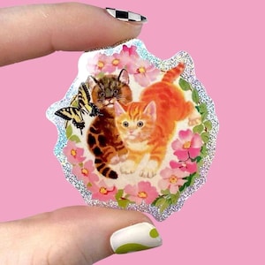 May include: A sticker featuring two kittens and a butterfly surrounded by pink flowers. One kitten is orange and white, the other is brown and tan. The sticker has a glittery, iridescent border.