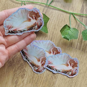 May include: A set of stickers featuring a classical painting of a woman reclining in a seashell. The stickers have a glittery, iridescent border. The woman has long, flowing hair and is surrounded by coral and rocks.