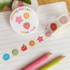 Könnte beinhalten: Washi-Tape-Rolle und runder Aufkleber mit farbenfrohem Design. Das Klebeband und der Aufkleber zeigen Illustrationen von Knöpfen, Sternen, Erdbeeren und Herzen. Der Text "Whimsy Buttons" und "Washi Tape" ist ebenfalls sichtbar.