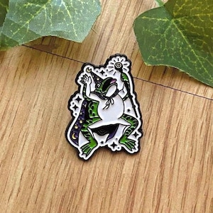 Könnte beinhalten: Ein skurriler Emaille-Pin mit einer Frosch-Zauberer-Darstellung. Der Frosch ist weiß mit grünen Akzenten und trägt einen Zaubererhut und -umhang mit Sternen- und Monddetails. Er hält einen Stab mit einem leuchtenden Orb. Der Pin hat eine schwarze Umrandung.