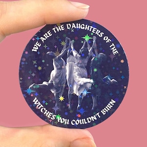 Könnte beinhalten: Ein runder Aufkleber mit dunkelblauem Hintergrund, holografischem Sternenmuster und einer schwarz-weißen Illustration von drei tanzenden Figuren. Der Text lautet: WE ARE THE DAUGHTERS OF THE WITCHES YOU COULDN'T BURN in weißer gotischer Schrift.