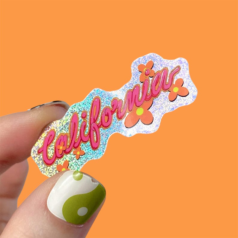May include: A holographic sticker with the word "California" in pink script, outlined in a darker pink. The sticker has orange flowers and a glittery background. The sticker is held up against an orange background.