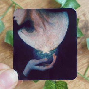 May include: A square sticker with an illustration of a figure with a glowing light emanating from their head, held in a hand. The artwork uses blue, white, and peach tones against a dark background. The image has a dreamlike quality.