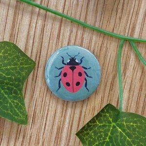 Könnte beinhalten: Ein runder Button mit hellblauem Hintergrund, der einen roten Marienkäfer mit schwarzen Punkten und Beinen zeigt. Der Marienkäfer ist auf dem Button zentriert. Grüne Blätter und ein Stiel umgeben den Button auf einer Holzoberfläche.