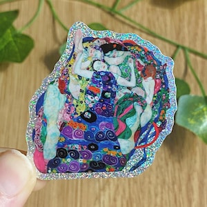 Op de afbeelding: Een sticker met een reproductie van Gustav Klimts "De Kus". Het kunstwerk toont omarmende figuren in levendige kleuren, waaronder goud, blauw, groen en rood, met een glinsterende rand. De sticker wordt door een hand vastgehouden.