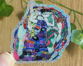 May include: A sticker featuring a colorful reproduction of Gustav Klimt's "The Kiss." The artwork depicts embracing figures in vibrant hues of blue, green, red, and gold, with a shimmering, glittery border. The sticker is held by a hand.
