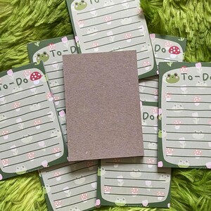 May include: A stack of green and white To-Do list notepads with a frog and mushroom design. The notepads have lined pages and the words "To-Do" printed at the top. A blank, brown notepad is in the center.
