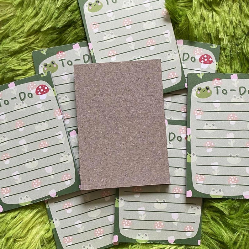 May include: A stack of green and white To-Do list notepads with a frog and mushroom design. The notepads have lined pages and the words "To-Do" printed at the top. A blank, brown notepad is in the center.
