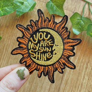 Op de afbeelding: Een ronde geborduurde patch met een gestileerd ontwerp van een zon en maansikkel in oranje, geel en zwart. Het midden bevat de gele geborduurde tekst "YOU ARE MY SUN SHINE" op een zwarte achtergrond met een maansikkelgezicht.