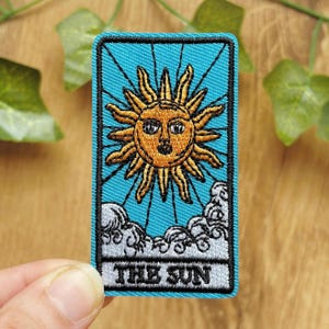 May include: A rectangular embroidered patch featuring a bright blue background with a central golden sun face surrounded by radiating rays. Below the sun, white clouds are depicted, and the text THE SUN is embroidered in black at the bottom.