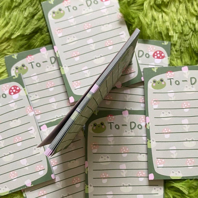 May include: A stack of To-Do list notepads with a green frog and mushroom design. Each notepad has a green border, lined paper, and the words "To-Do" at the top. The background is a green, fuzzy surface.