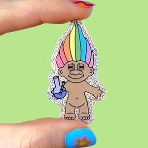 May include: A glittery sticker featuring a cartoon troll with rainbow hair holding a blue bong. The troll has a smiling face and is tan-skinned. The sticker has a silver glitter border and is held against a light green background.