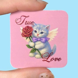 Könnte beinhalten: Ein rosa Quadrat mit einer Illustration eines hellblauen Kätzchens. Das Kätzchen hat Engelsflügel, eine lila Schleife und hält eine rote Rose. Glitzernder Schriftzug "True Love".