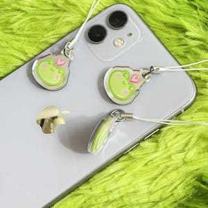 May include: Clear acrylic frog charms with a white string attached. The frog charms have a green body, pink cheeks, and a pink heart. The charms are on a silver phone.