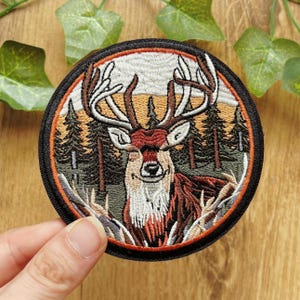 Könnte beinhalten: Ein runder, bestickter Aufnäher mit einem detaillierten Hirsch mit großem Geweih vor einer Waldlandschaft mit Tannen. Das Design verwendet braune, beige, grüne und weiße Fäden und ist von einem dicken schwarzen Rand mit einem orangefarbenen Innenring umgeben.