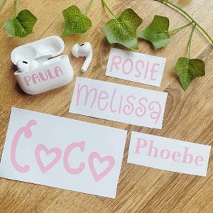 Könnte beinhalten: Weiße kabellose Ohrhörer und Ladecase mit dem Namen "Paula" in rosa Schrift. Außerdem rosa Namensaufkleber für "Rosie", "Melissa", "Coco" und "Phoebe". Grüne Efeublätter befinden sich im Hintergrund.