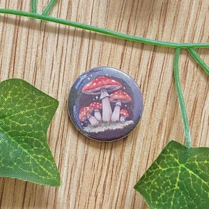 Peut inclure: Un bouton rond représentant un groupe de champignons à chapeaux rouges et tiges blanches sur fond bleu foncé. Le bouton a une finition brillante, entouré de feuilles vertes et d'une surface en bois.