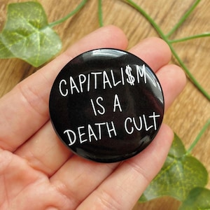 Könnte beinhalten: Ein runder, schwarzer Ansteckbutton mit weißem, handgeschriebenem Text, der CAPITALI$M IS A DEATH CULT lautet. Das Design verwendet ein Dollarzeichen anstelle des Buchstabens S im Wort capitalism und bietet einen klaren Kontrast.