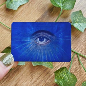 Può includere: Una carta rettangolare blu con un occhio dipinto al centro. Linee azzurre irradiano dall'occhio su uno sfondo blu più scuro. La carta è tenuta in mano, con foglie verdi sullo sfondo.