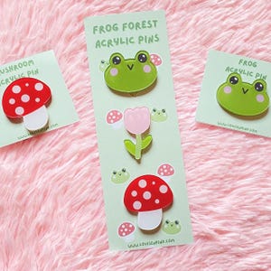 Può includere: Spille in acrilico con disegni di funghi, tulipani e rane. Le spille sono su biglietti verde chiaro con il testo "Frog Forest Acrylic Pins" e "Mushroom Acrylic Pin". Lo sfondo è una superficie morbida e rosa.