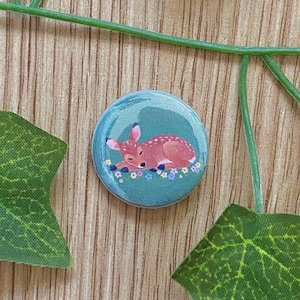 Könnte beinhalten: Ein runder Button mit türkisblauem Hintergrund, der eine Illustration eines schlafenden Rehs zeigt. Das Reh ist braun mit weißen Flecken und liegt auf kleinen blauen und weißen Blumen. Grüne Blätter und eine grüne Ranke umgeben den Button.