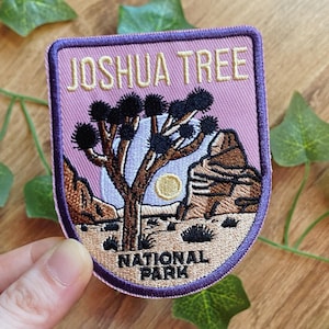 Könnte beinhalten: Gestickter Aufnäher mit einem Design des Joshua Tree Nationalparks. Der Aufnäher hat einen lila Rand und einen hellvioletten Hintergrund mit einem Joshua-Baum, einer Sonne und Felsformationen. Der Text "JOSHUA TREE NATIONAL PARK" ist auf den Aufnäher gestickt.