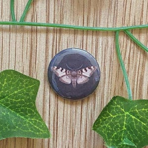 Op de afbeelding: Een ronde, donkergrijze knoop met een gedetailleerde illustratie van een mot met crème- en bruine vleugels. De mot is gecentreerd op de knoop, die wordt omgeven door groene bladeren en een groene wijnstok tegen een achtergrond van houtnerf.