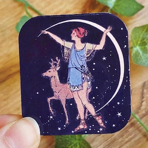 May include: A square sticker featuring a celestial design. It depicts a woman in a blue dress, a deer, and a crescent moon against a dark blue starry background. The woman is holding a wand, and the deer is standing beside her.
